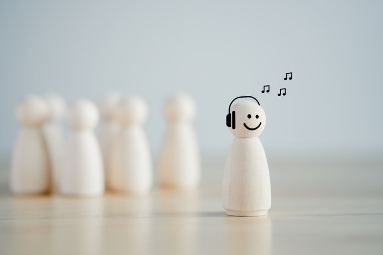 A smiling wooden peg figure with headphones stands out from a line of plain peg figures in the background, for the Social Jam blog post on how introvert business owners can shine online.